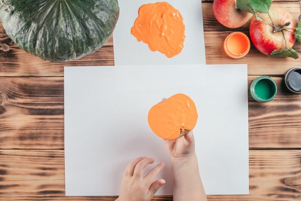 Nietradycyjne techniki rysowania improwizowanymi środkami Step-by-step-halloween-childrens-tutorial-pumpkin-apple-prints-step-7-child-hand-holds-half-apple-painted-orange-gouache-make-print-top-view