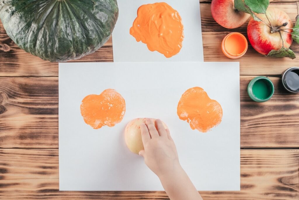 Nietradycyjne techniki rysowania improwizowanymi środkami Step-by-step-halloween-childrens-tutorial-pumpkin-apple-prints-step-8-child-hand-leaves-prints-paper-with-half-apple-painted-orange-gouache-paint-top-view